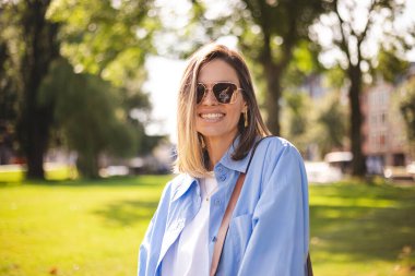 Güneş gözlüklü, tişörtlü, gülümseyen ve kırsal kesimde kameraya bakan güzel bir kadın. Açık havada gülümseyen sarışın bir kızın portresi. Mutlu neşeli kız yeşil şehre gülüyor, modaya bak..