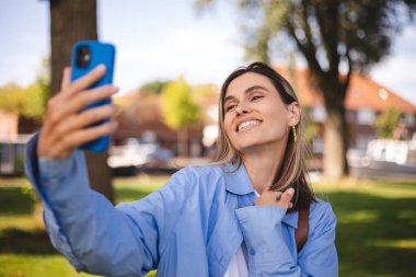Güzel sarışın kadın kamerada selfie çekiyor. Tatlı kız parkta yürüyor ve kameraya ya da aynaya bakıyor. Doğada, ince kız kamerayla flört eder, gülümser ve güler, video çağrısı.