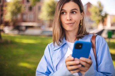 Elinde telefon olan bir kadın. Dışardan düşünen şaşkın bir kadın. Akıllı telefonu olan şüpheli bir kadın. Bilgi okumayı, arama çözümü bulmayı düşün. Kız gömlek, çanta, bluz giyer, parkta yürür.