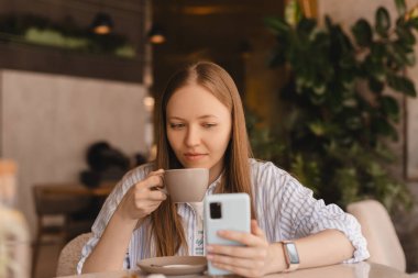 Genç ve güzel bir kadın cep telefonunu tutuyor ve kafeteryada otururken akıllı telefona bakıyor. Mutlu kız cep telefonu kullanıyor ve flört ediyor. Kahve içen, gülümseyen, kafede oturan bir kadın..