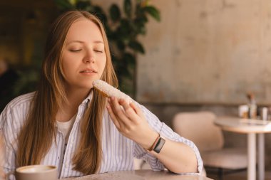 Genç sarışın kadın kafede oturmuş ekkin yiyor. Dişi kruvasan parçası restoranda neşeli görünüyor. Yemek günü konseptini aldatmak. Kadın ekkin yemek için iştahla hazırlanıyor. Hindistan cevizi fırınının tadını çıkar..