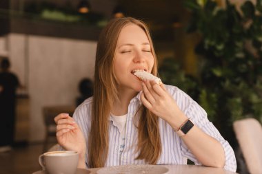 Genç sarışın kadın kafede oturmuş ekkin yiyor. Dişi kruvasan parçası restoranda neşeli görünüyor. Yemek günü konseptini aldatmak. Kadın ekkin yemek için iştahla hazırlanıyor. Hindistan cevizi fırınının tadını çıkar..