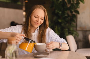 Sarışın kadın masada oturmuş, restoranda çay içiyor. Kadın kendisi ve cam çaydanlıktan arkadaşı için portakal çayı döken bir gömlek giyer ve kafede fincanlara doldurur. Mutlu kız ve arkadaşlarınla dinlen, kokunun tadını çıkar..