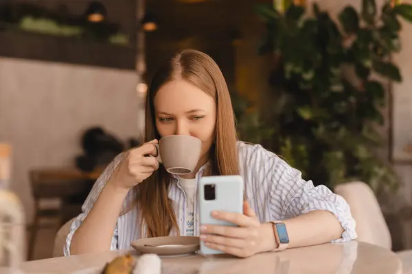 Genç ve güzel bir kadın cep telefonunu tutuyor ve kafeteryada otururken akıllı telefona bakıyor. Mutlu kız cep telefonu kullanıyor ve flört ediyor. Kahve içen, gülümseyen, kafede oturan bir kadın..