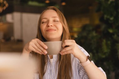 Güzel, gülümseyen, kahve kokan ve restoranda dinlenirken fincandan kapuçino içen sarışın kadının portresi. Kadın soyunmuş gömlek giyiyor..