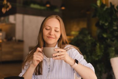Güzel, gülümseyen, kahve kokan ve restoranda dinlenirken fincandan kapuçino içen sarışın kadının portresi. Kadın soyunmuş gömlek giyiyor..