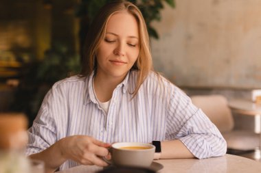 Güzel, gülümseyen, kahve kokan ve restoranda dinlenirken fincandan kapuçino içen sarışın kadının portresi. Kadın soyunmuş gömlek giyiyor..