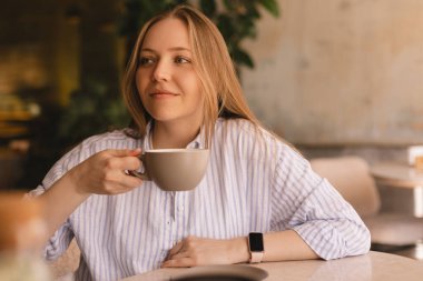 Güzel, gülümseyen, kahve kokan ve restoranda dinlenirken fincandan kapuçino içen sarışın kadının portresi. Kadın soyunmuş gömlek giyiyor..