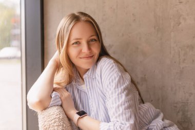 Evdeki portre için poz veren sarışın kadın portresi, rahat koltukta oturuyor, gülümsüyor, gülüyor, başını yana yaslıyor. Şehvetli görün ve rüya gör. Gömleğini giy.