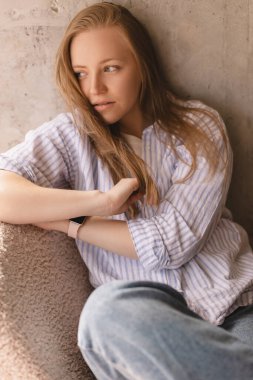 Evinde portre için poz veren sarışın kadın portresi, rahat koltukta oturuyor, gülümsüyor, gülüyor, saçını düzeltiyor. Kız şehvetli görünüyor ve rüya görüyor. Gömlek ve kot pantolon giyiyor..