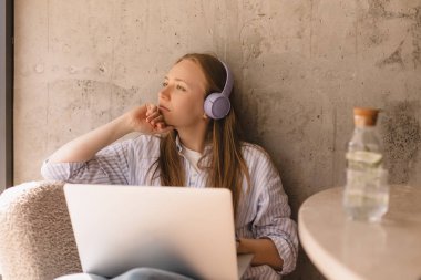 Kafede dizüstü bilgisayarla çalışırken gülümseyen, neşeli, genç sarışın kadın çalışıyor ve dinleniyor, düşünceli ve hayalperest görünüyor. Kablosuz kulaklıklı ya da kulaklıklı bir kız müzik ya da podcast dinliyor..