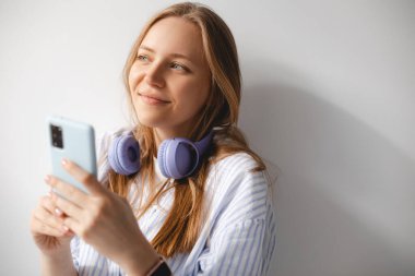 Düşünceli genç bir kadın beyaz tişörtlü, ensesinde kablosuz mor kulaklıklı. Uygulama, sosyal medya, bağımlılık, ağ konsepti. Hayalperest kız cep telefonunu beyaz duvarda tutuyor.