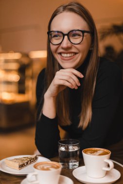 Kafede arkadaşlarıyla oturup gülen mutlu bir kadın. Güzel konuşmaydı. Gülen kız elinde kek, iki fincan kahve ve masada bir bardak suyla oturuyor. Sarışın kadın gözlük ve siyah kazak giyiyor..