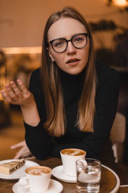 Attractive young blonde woman attentively listen, look at camera, she waiting for answer. Girl in eye glasses sitting in cafe, look questioningly, spread hand. What, question. clipart