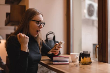Neşeli sarışın kadın cep telefonundan bir iş mesajı aldı, iyi haberler okuyordu, piyangoyu kazanmayı kutluyordu, kazanan jest yaparken çok mutlu ve neşeli hissediyordu, kahve fincanıyla masada oturuyordu..
