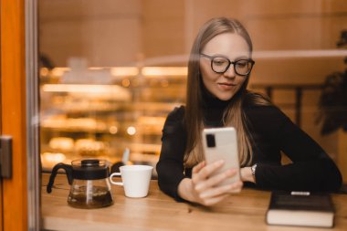 Happy, gözlüklü hoş bir bayanın selfie çekmesinden ya da telefon görüşmesinden hoşlanırdı. Kafe arka planında izole bir masada otururdu. Mutlu kadın gülümsüyor ve cep telefonundaki selfie kamerasıyla fotoğraf çekiyor..