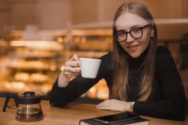 Güzel sarışın kız bir kafede çay içiyor. Bir restoranda masada oturmuş çay fincanına hoş geldin diyen güzel bir kız. Kız bardağı tut ve gülümseyen kameraya bak..