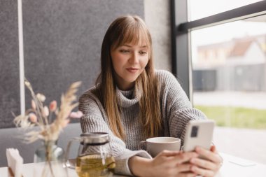 Genç gülümseyen sarışın kızın portresi akıllı telefondan en iyi arkadaşına video araması yapıyor, ara veriyor, iyi haberler veriyor, kafede oturuyor, selfie çekiyor. Kadınlar gri kazak giyer.