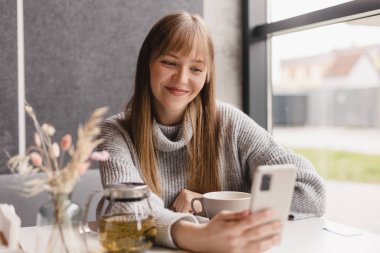 Genç gülümseyen sarışın kızın portresi akıllı telefondan en iyi arkadaşına video araması yapıyor, ara veriyor, iyi haberler veriyor, kafede oturuyor, selfie çekiyor. Kadınlar gri kazak giyer.