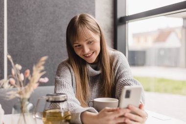Genç gülümseyen sarışın kızın portresi akıllı telefondan en iyi arkadaşına video araması yapıyor, ara veriyor, iyi haberler veriyor, kafede oturuyor, selfie çekiyor. Kadınlar gri kazak giyer.