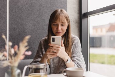 Genç ve güzel bir kadın cep telefonunu tutuyor ve kafeteryada otururken akıllı telefona bakıyor. Mutlu kız cep telefonu kullanıyor ve flört ediyor. Çay içen, gülümseyen, kafede oturan bir kadın..