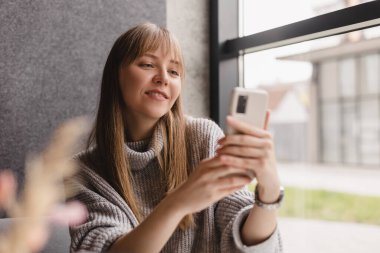 Genç ve güzel bir kadın cep telefonunu tutuyor ve kafeteryada otururken akıllı telefona bakıyor. Mutlu kız cep telefonu kullanıyor ve flört ediyor. Çay içen, gülümseyen, kafede oturan bir kadın..