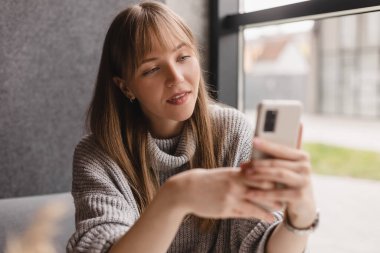 Genç ve güzel bir kadın cep telefonunu tutuyor ve kafeteryada otururken akıllı telefona bakıyor. Mutlu kız cep telefonu kullanıyor ve flört ediyor. Çay içen, gülümseyen, kafede oturan bir kadın..