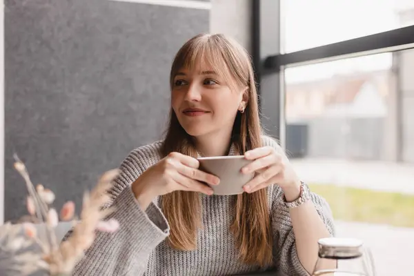 Lokantada çay ve aromanın tadını çıkaran neşeli genç bir kadının portresi. Gülümseyen güzel kız kafede sıcak çay içiyor. Gri kazak giyip kahkahalar atan heyecanlı sarışın kadın..