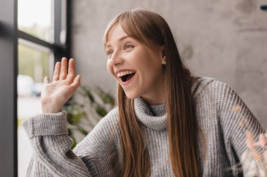 Çekici, neşeli, yetişkin bir kadın, arkadaşına merhaba demek için el sallıyor, kafe restoranında yürüyor, kadın, kafedeki arkadaşını rahatça selamlıyor..
