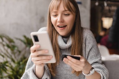 Müşteri kartı tutan mutlu genç sarışın kadın cep telefonu uygulaması kullanıyor. Moda kıyafetleri alıyor, internetten alışveriş yapıyor, akıllı telefondaki ecommerce dijital mağazasından alışveriş yapıyor..