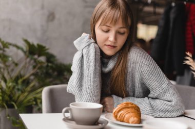 Kafede otururken kruvasan yiyen genç bir kadın, çay molası. Güzel ve aç bir kadın zevkle lezzetli kruvasan yiyor. Sarışın kadın öğle yemeğinin tadını çıkar, gri kazak giy..