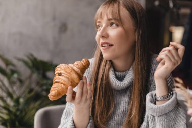 Kafede otururken kruvasan yiyen genç bir kadın, çay molası. Güzel ve aç bir kadın zevkle lezzetli kruvasan yiyor. Sarışın kadın öğle yemeğinin tadını çıkar, gri kazak giy..