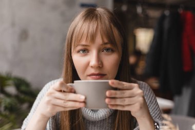 Sabahları bir fincan çay içen mutlu genç bir kadın. Bir fincan sıcak çayla restoranda oturan hayalperest kız, kameraya bak. Kafede gri kazak giyen güzel bir kadın. İyi günler, dinlenme.