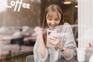 Cep telefonuyla video oyunu oynayan neşeli sarışın kadın, iyi haberler okuyor, piyangoyu kazanmayı kutluyor, kazanan hareketi yaparken çok mutlu ve neşeli hissediyor, cam camların altındaki masada oturuyor..