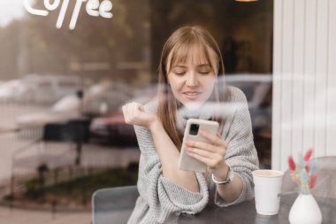 Cep telefonuyla video oyunu oynayan neşeli sarışın kadın, iyi haberler okuyor, piyangoyu kazanmayı kutluyor, kazanan hareketi yaparken çok mutlu ve neşeli hissediyor, cam camların altındaki masada oturuyor..