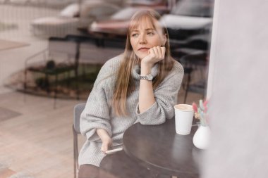 Telefon kullanan genç, güzel bir kadın akıllı telefonlu bir kafede oturuyor, mesajlara cevap veriyor, telefon ediyor, mektuplar yazıyor, pencerenin altına fotoğraf koyuyor. Kadın rüya görüyor, cevap vermeyi düşün, gri kazak giy.