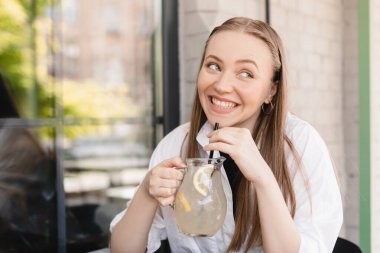 Sıcak bir yaz gününde bir bardak limonatayla çekici bir genç kadın. Güzel dişi hayal kuruyor ve mutlu görünüyor, gülümsüyor. Yaz havası, taze içkiler. Dışarıdaki kafede limonatayla oturan kız..