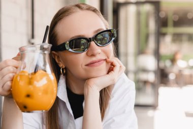 Gülümseyen beyaz kadın kameraya bakıyor açık kafede kokteyl içiyor. Sarışın kadın siyah gözlük takıyor ve güneşli havadan hoşlanıyor. Restoranın dışında yaz tatili konsepti. Şerefe..