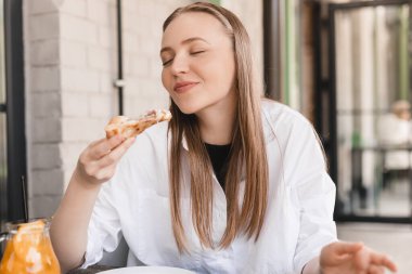 Sokak kafesinde lezzetli pizzalar yiyen bir kadın. Güneşli bir günde fast food servisi. Sarışın kadın, restoran masasında beyaz gömlek, turuncu limonata ve pizza giyiyor. Lezzetli pizzadan hoşlanan bir kadın..