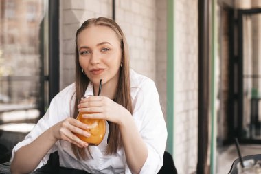 Kafeteryada portakallı limonata içen beyaz bir kadının yakın çekimi. Beyaz gömlekli sarışın susuzluğunu gideriyor. Rahatlama konsepti. Mutlu gülen kadın.