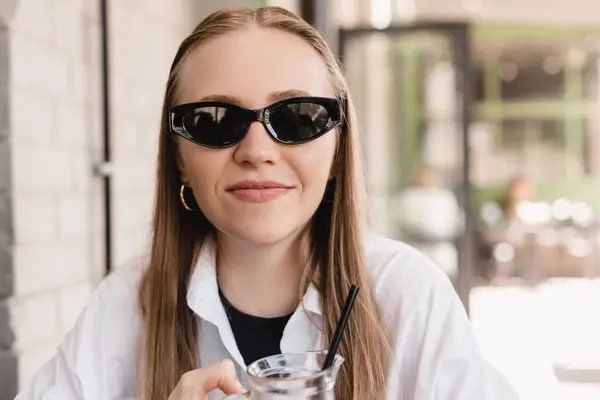 Gülümseyen beyaz kadın kameraya bakıyor açık kafede kokteyl içiyor. Sarışın kadın siyah gözlük takıyor ve güneşli havadan hoşlanıyor. Restoranın dışında yaz tatili konsepti. Şerefe..