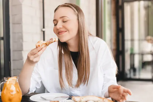 Sokak kafesinde lezzetli pizzalar yiyen bir kadın. Güneşli bir günde fast food servisi. Sarışın kadın, restoran masasında beyaz gömlek, turuncu limonata ve pizza giyiyor. Lezzetli pizzadan hoşlanan bir kadın..