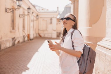 Genç sarışın kadın sokakta cep telefonu kullanıyor. Eski şehrin caddesinde duran kız akıllı telefondaki bilgilere bakıyor, arkanı dön ve kameraya bak..