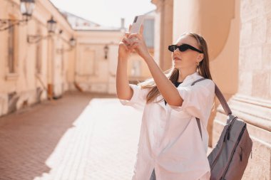 Sırt çantalı güzel sarışın kadın turist manastır ya da kilise yakınlarındaki eski şehir caddesinde yürürken cep telefonuyla fotoğraf çekiyor. Tarihi yerlerde yalnız yürüyen bir kız..