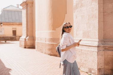 Genç sarışın kadın sokakta cep telefonu kullanıyor. Eski şehrin caddesinde duran kız akıllı telefondaki bilgilere bakıyor, arkanı dön ve kameraya bak..