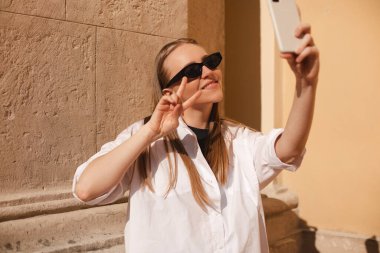 Sırt çantalı güzel, sarışın bir turist. Eski şehir caddesinde yürürken selfie çekiyor ve barış jesti yapıyor. Kadın siyah gözlük, beyaz gömlek ve gri sırt çantası takıyor..