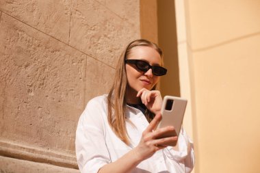 Genç sarışın kadın sokakta cep telefonu kullanıyor. Eski şehrin caddesinde duran kız akıllı telefondaki bilgilere bakıyor, arkanı dön ve kameraya bak..