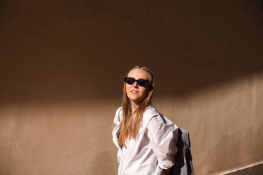 Eski şehirde bej duvarın yanında duran çekici bir kadın. Güneş ışığının kızı, siyah güneş gözlüğü tak ve beyaz gömlek giy güneşe bak. Güneşli yazın gölgesi ve ışığı. 