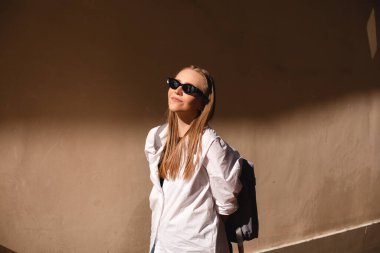 Eski şehirde bej duvarın yanında duran çekici bir kadın. Güneş ışığının kızı, siyah güneş gözlüğü tak ve beyaz gömlek giy güneşe bak. Güneşli yazın gölgesi ve ışığı. 