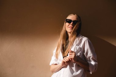 Eski şehirde bej duvarın yanında duran çekici bir kadın. Güneş ışığının kızı, siyah güneş gözlüğü tak ve beyaz gömlek giy güneşe bak. Güneşli yazın gölgesi ve ışığı. Kadın saçını düzeltiyor.. 